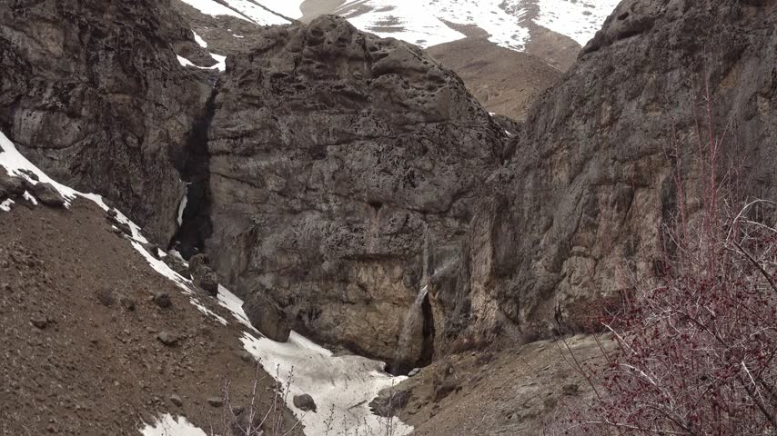 A narrow stream of water flows over a steep mountain gorge wall into a basin filled with remnants of seasonal snow and arid brown earth terrain