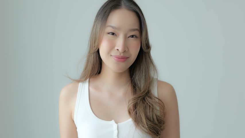 Young Asian adult woman portrait wearing white tank top, smiling with thumbs up in soft studio light