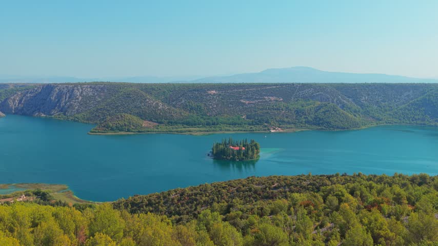 Drone shot captures Visovac Monastery on a small island, encircled by clear blue lake waters and lush hills under bright daylight, perfect for travel, nature, and heritage themes.