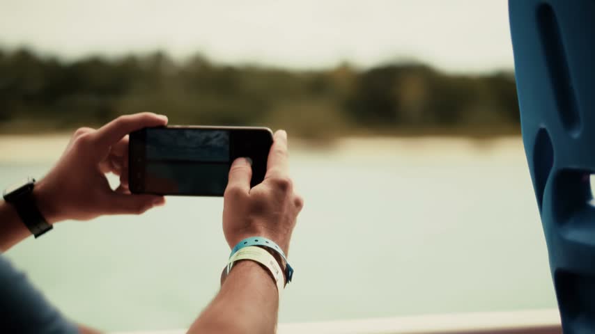 Tourist holding smartphone in outdoor setting. This smartphone captures landscape with scenic view. Photo taking records nature scene on water surface. Hand holding device creates photo for travel memory. Landscape photography shows beach and green trees. Smartphone camera records photo in bright daylight. Tourist capturing landscape on sunny summer day