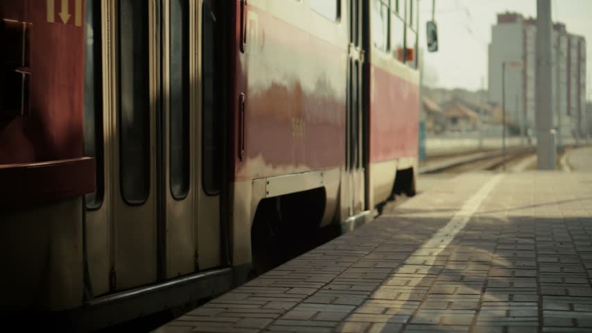 Tram moving on railway track in city transport. This tram arrives at station platform with route display. Urban transport stopping near metal fence in morning light. Tram track leads through residential block and apartment building. City tram carries passengers in public transport network.  Station platform connects commuter route in urban landscape