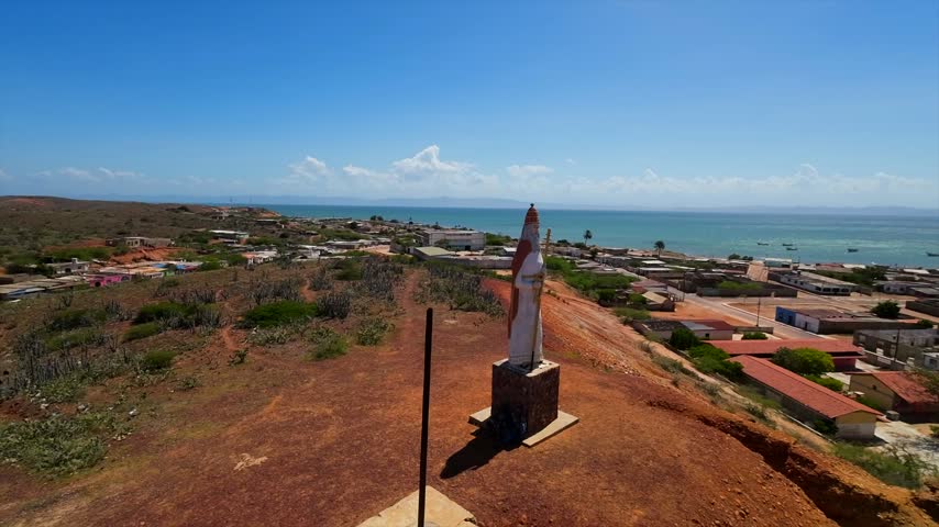 Aerial view circling a statue on a hill above a coastal fishing town and beach