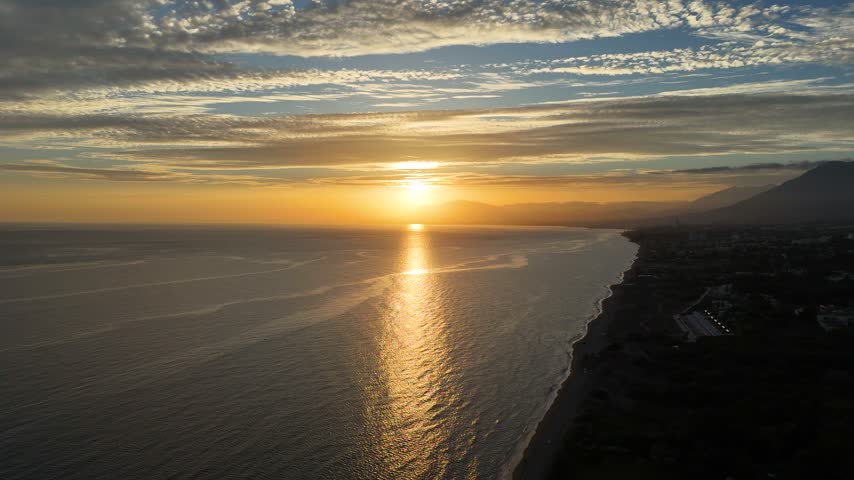 Drone aerial view of the sun setting directly on the horizon over the Elviria coastline near Marbella, Costa del Sol, Spain. Golden light reflects across the calm Mediterranean Sea, with silhouetted mountains and the coastal town fading into a soft, hazy evening sky above the quiet shoreline, where the coast fades into twilight.