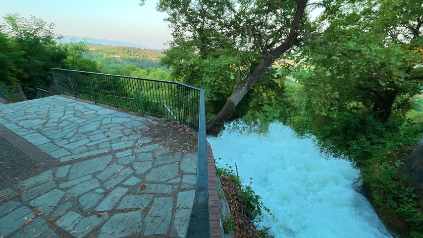 Rugged stone path hugs steep, verdant cliffside. Whitewater crashes below, frothy and wild. Green foliage sways with wind, framing the scene. Location: Edessa waterfalls. Balkans, Greece