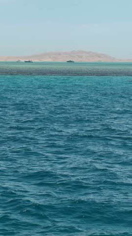 Waves splash gently in the blue waters of Hurghada, Egypt. Boats move near the distant islands under a clear sky.