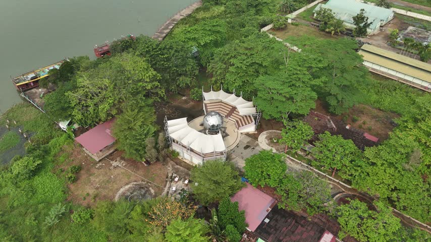 An aerial view of a riverside amphitheater in Pontianak, featuring a white-roofed stage surrounded by lush trees, walking paths, and buildings along the riverbank.