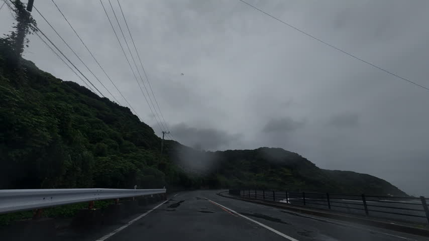 Smooth 4K 24p forward vehicle POV shot navigating a sweeping curve on a rural Japanese highway through lush mountains under a heavy overcast sky. Authentic raindrops striking the windshield and lens with crisp wet pavement reflections. Minimalist cold desaturated Rec.709 color grade. Excellent establishing shot for travel videos, mood transitions, mindfulness visual loops, and automotive commercials.