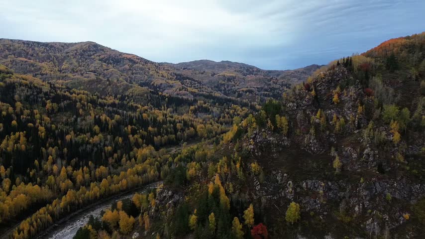 Aerial view of a mountainous forest landscape with rocky hills, winding roads and dense golden autumn foliage under a partly cloudy sky