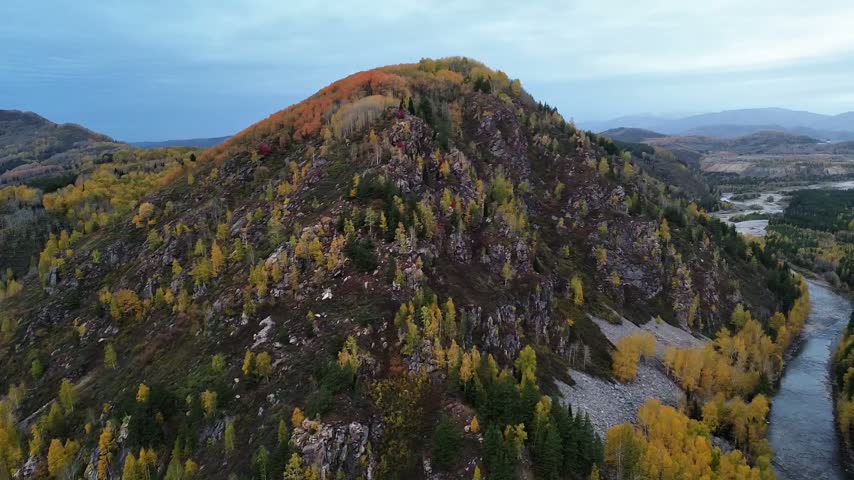 Aerial drone view of a rocky mountain valley with a winding river, forested slopes, and golden autumn foliage covering the surrounding hills