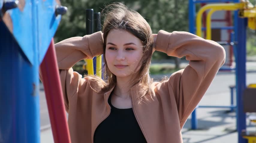 Portrait of a beautiful teenage student standing with her hands behind her head, relaxing in a city park playground on a sunny day as the wind blows through her hair