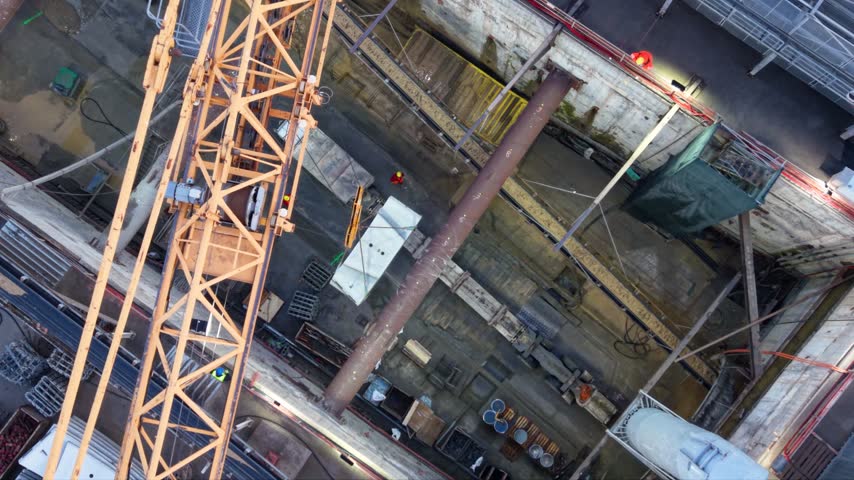 Top down aerial rotation above a deep braced construction excavation crossed by a tower crane boom, with workers, large steel supports and equipment below.