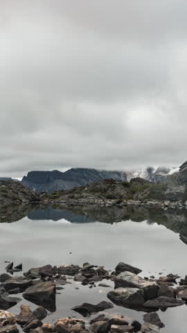 A vertical timelapse captures clouds moving over snow-capped mountains reflected in a calm alpine lake with rocky shoreline.