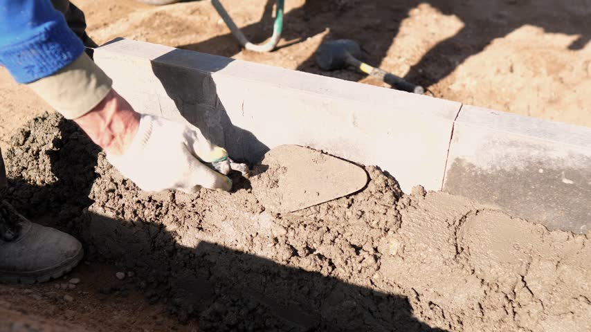 Construction worker smooths fresh concrete mortar along block curb at outdoor building site, using hand trowel in bright sunlight with dirt ground, boots, and unfinished wall.