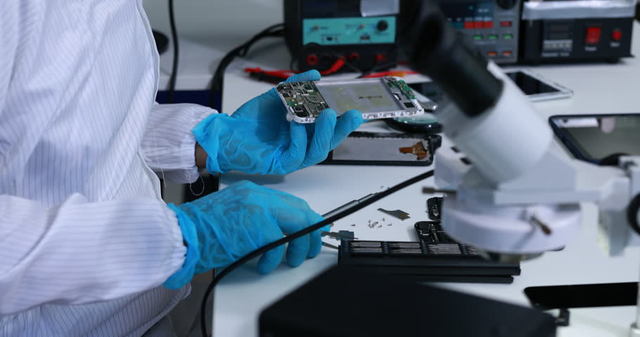 Technician wearing blue gloves repairs a smartphone circuit board at a laboratory workbench with microscope and electronic testing equipment.