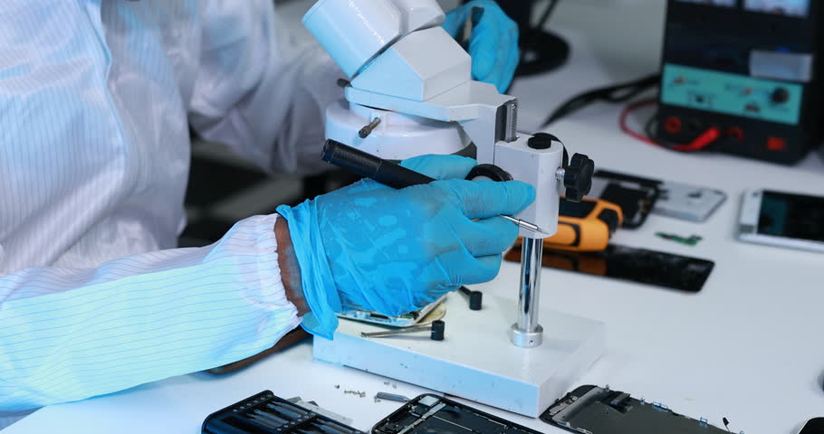 Technician in protective gloves repairing a smartphone circuit board under a microscope at an electronics workbench.