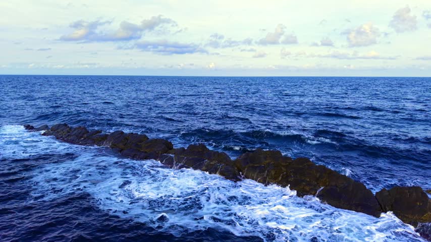 View of ocean waves crashing against rocky shoreline near coastline. Waves crash against rocks along a coastline under a cloudy sky during late afternoon.