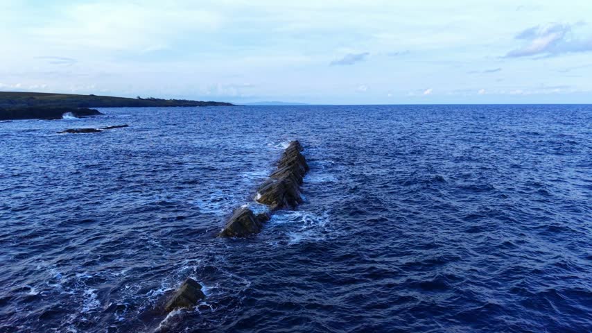Rocky coastline meeting the ocean under a blue sky at a remote location. Rocky formations extend into the ocean with waves crashing against them on a clear day.