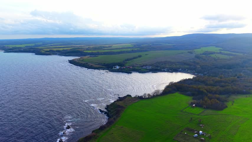 Scenic view of a shoreline and green fields under a cloudy sky. The scene shows a coastline meeting green fields with patches of grass under cloudy skies.