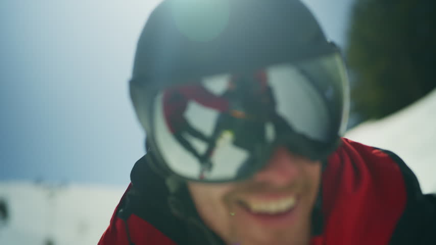A man wearing a ski helmet and goggles smiles on a sunny winter day. His goggles reflect friends skiing down the snowy mountain.