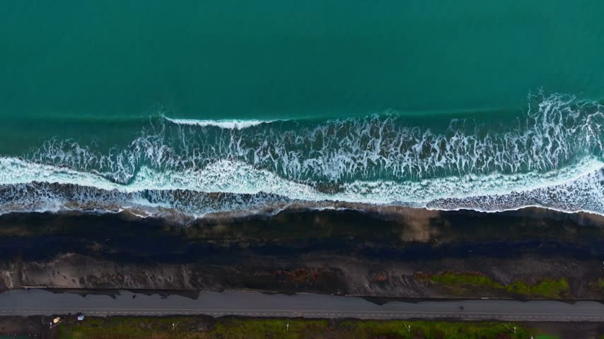Waves crash onto a rocky shore near a road under an open sky. Waves crash onto dark rocks along a shoreline near a road. The scene shows the ocean meeting land.