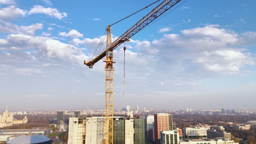 Aerial view of a large tower crane above a high-rise construction site in Bucharest, Romania, with modern office buildings, busy urban roads, residential districts and green parkland stretching across the city.