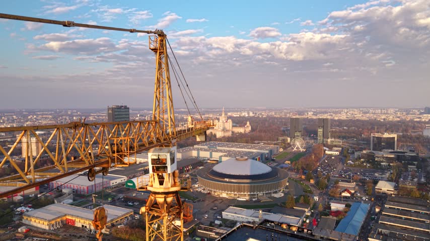 Aerial view of a large tower crane above a high-rise construction site in Bucharest, Romania, with modern office buildings, busy urban roads, residential districts and green parkland stretching across the city.