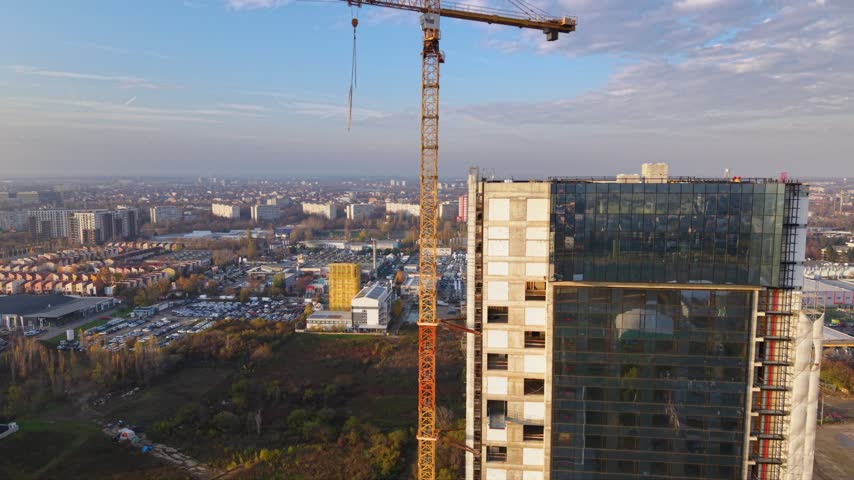Aerial view of a large tower crane above a high-rise construction site in Bucharest, Romania, with modern office buildings, busy urban roads, residential districts and green parkland stretching across the city.