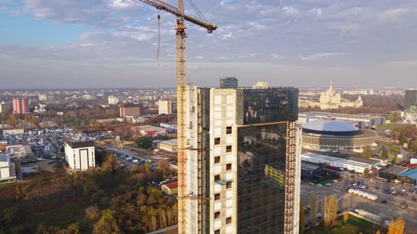 Aerial view of a large tower crane above a high-rise construction site in Bucharest, Romania, with modern office buildings, busy urban roads, residential districts and green parkland stretching across the city.