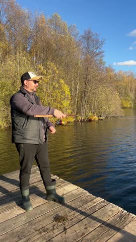 A middle-aged Caucasian man with short hair stands on a wooden dock, fishing in a serene lake surrounded by autumn foliage.