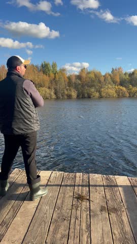 A man stands on a wooden dock by a lake, fishing in autumn. The scene features colorful trees reflecting in the water under a clear blue sky.