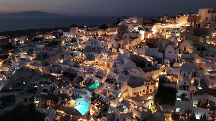 Golden lights sparkle across the traditional white buildings of Oia in Santorini as dusk settles over the Aegean Sea and island coastline.