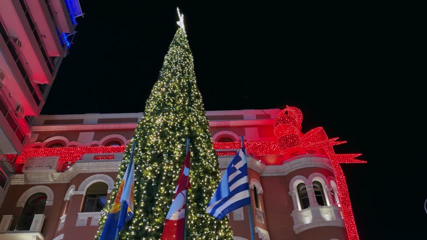 Thessaloniki city center glows with a brightly lit Christmas tree and building decorations under a dark night sky. Capturing the authentic festive atmosphere of the Greek holidays.