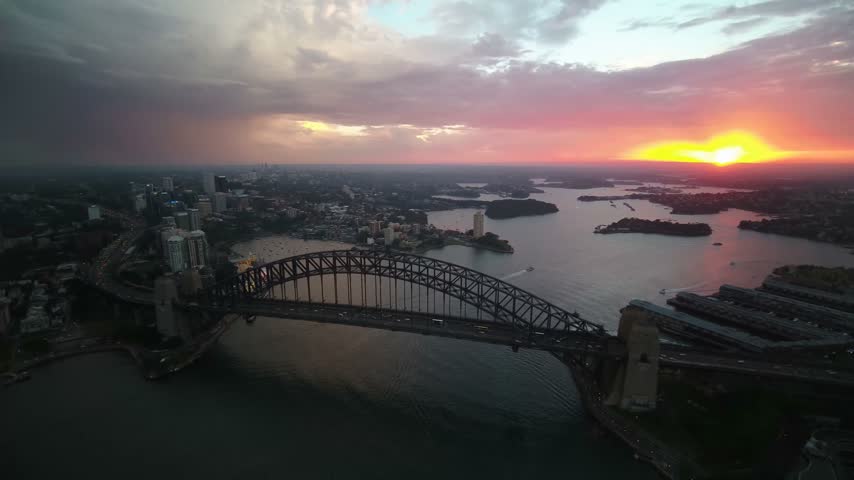 High-angle aerial drone perspective looking west over the massive steel arch of the Sydney Harbour Bridge, capturing a vibrant orange sunset over the Parramatta River, local harbor islands, and distant suburban shorelines under dramatic twilight clouds in Sydney, New South Wales, Australia.