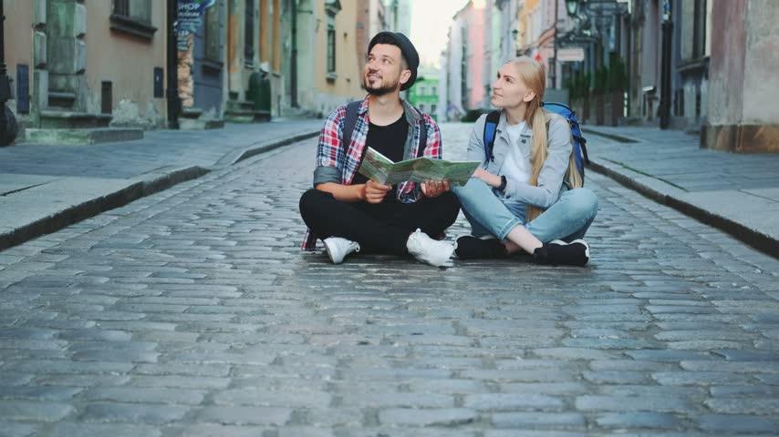 A young man and woman sit cross-legged on a cobblestone street in a historic city center, using a large paper map to navigate during daytime travel.