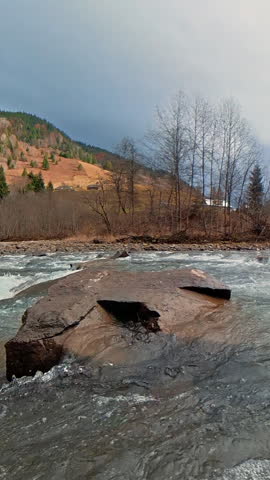Fast mountain river flowing around a large rock in autumn season, Wide scenic view of powerful rapids beneath forested slopes and changing clouds