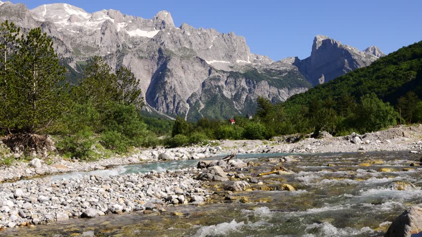 Panoramic mountain landscape in Albania, showcasing rugged peaks, green valleys, rocky slopes, and vast natural scenery under an expansive sky. Layers of mountains stretch toward the horizon, creating a dramatic sense of depth and wilderness. Sunlight moves across the terrain, revealing contrasting textures of forests, grasslands, exposed rock, and distant ridges. The remote setting reflects the untouched beauty of the Albanian countryside and the diverse character of the Balkan landscape. Ideal footage for travel documentaries, tourism campaigns, hiking content, nature films, outdoor adventure productions, environmental projects, and cinematic landscape sequences. The spectacular panorama conveys freedom, exploration, tranquility, wilderness, and the enduring appeal of European mountain destinations.