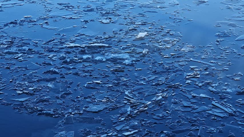Close-up of spring thaw on reservoir. Small pieces of broken ice on smooth surface of blue water. River current carries thin layer of melting ice, winter, early spring. Natural background with ice