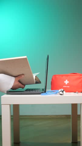 Vertical video doctor hand holding a medical textbook while working on a laptop, with a stethoscope and first aid kit nearby, representing healthcare education and telemedicine