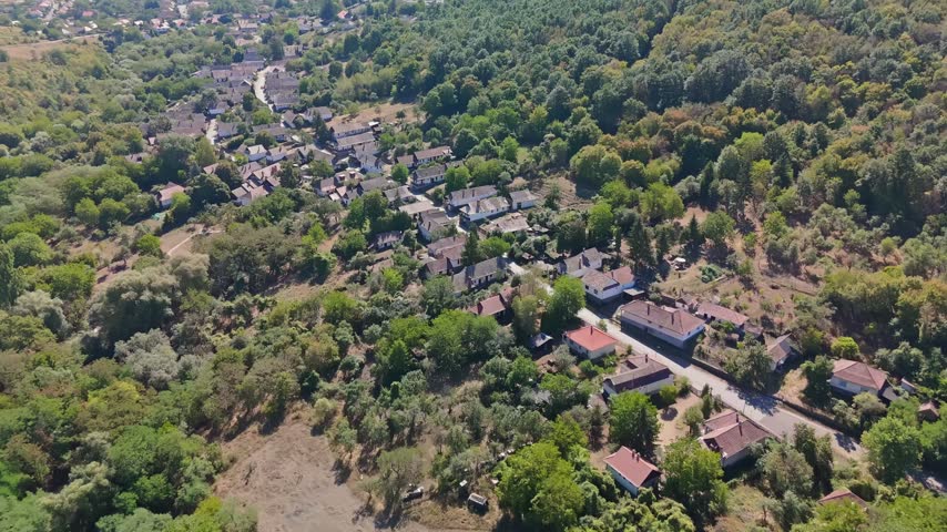 Aerial view of traditional village houses and gardens in Hollókő, Hungary, surrounded by wooded Cserhát hills, rural roads, and surrounding summer greenery.