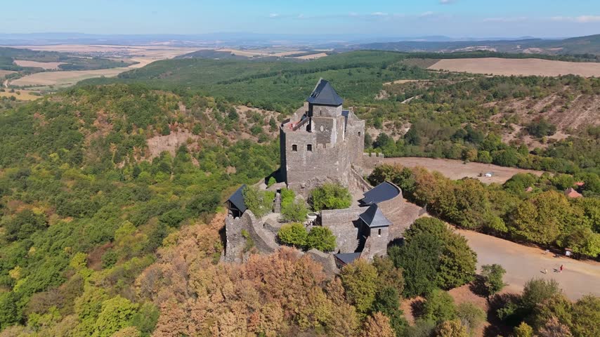 Aerial view of medieval Hollókő Castle in Hungary, with stone walls and tower above late summer woodland and patchwork farmland in the Cserhát Mountains.
