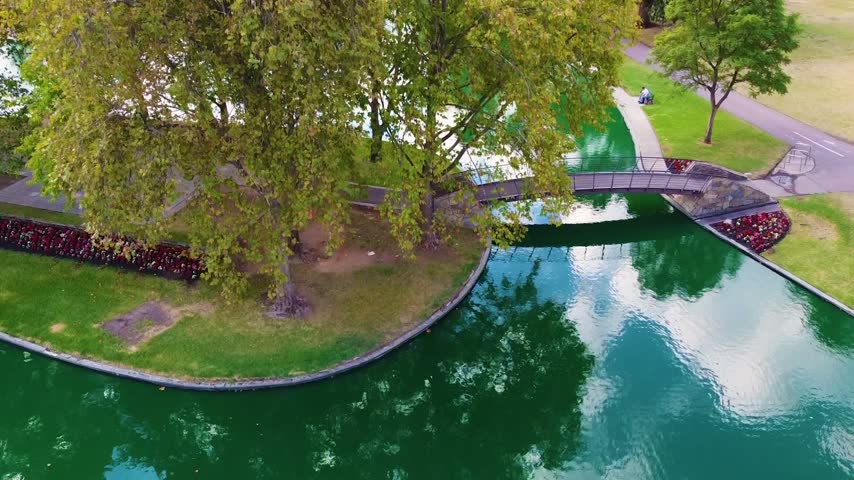 Aerial footage of a tranquil park with a winding river, lush trees, and a pedestrian bridge