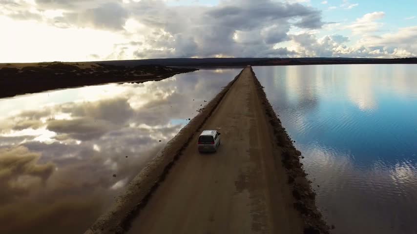 Aerial view following a van driving along a narrow dirt causeway separating two lakes