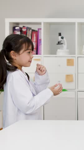 Young elementary school girl wearing white lab coat playfully acts out game in science classroom.