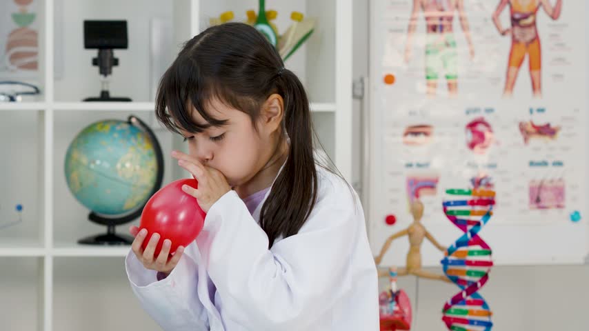 Young elementary student in a white lab coat inflates a red balloon during science class.