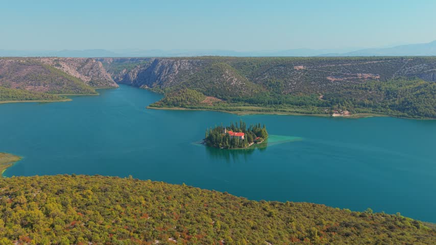 Drone closing into Visovac Monastery on a forested island, surrounded by turquoise lake waters and rugged hills in bright daylight, showcasing tranquil scenery.