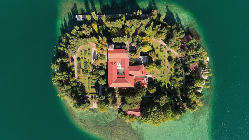 Top down drone shot of Visovac Monastery atop a lush green island encircled by vibrant turquoise lake waters, captured from directly above in crisp sunlight and vivid color.