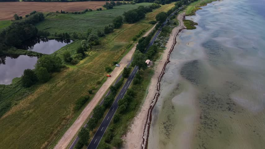 Coastal road lined with leafy trees runs between green farmland and cloudy turquoise waters along sandy shoreline near small inland pond and parked car, German Baltic Sea. Aerial drone wide shot