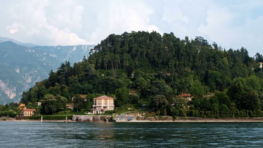 Panning shot of beautiful historic villas and luxury hotels on waterfront of Lake Como under blue sky in summer.