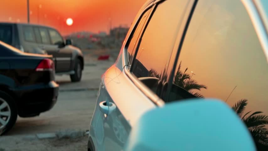Close up of parked cars in an outdoor lot during a golden hour sunset. A cinematic shallow depth of field highlights a silver vehicle showing palm tree reflections on its window glass.