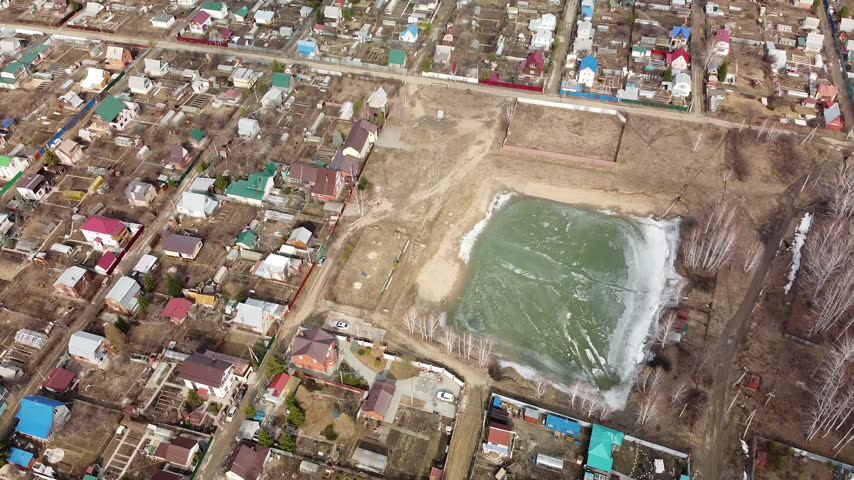 Aerial view of icy pond surrounded by residential homes. Greenish water frozen solid, edged in snow. Neighborhood houses dot the landscape nearby. Use for construction, winter scenes